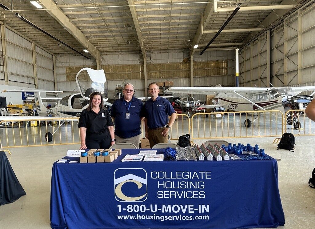 Three people standing at the Collegiate Housing Services desk in the LIFT Academy hangar